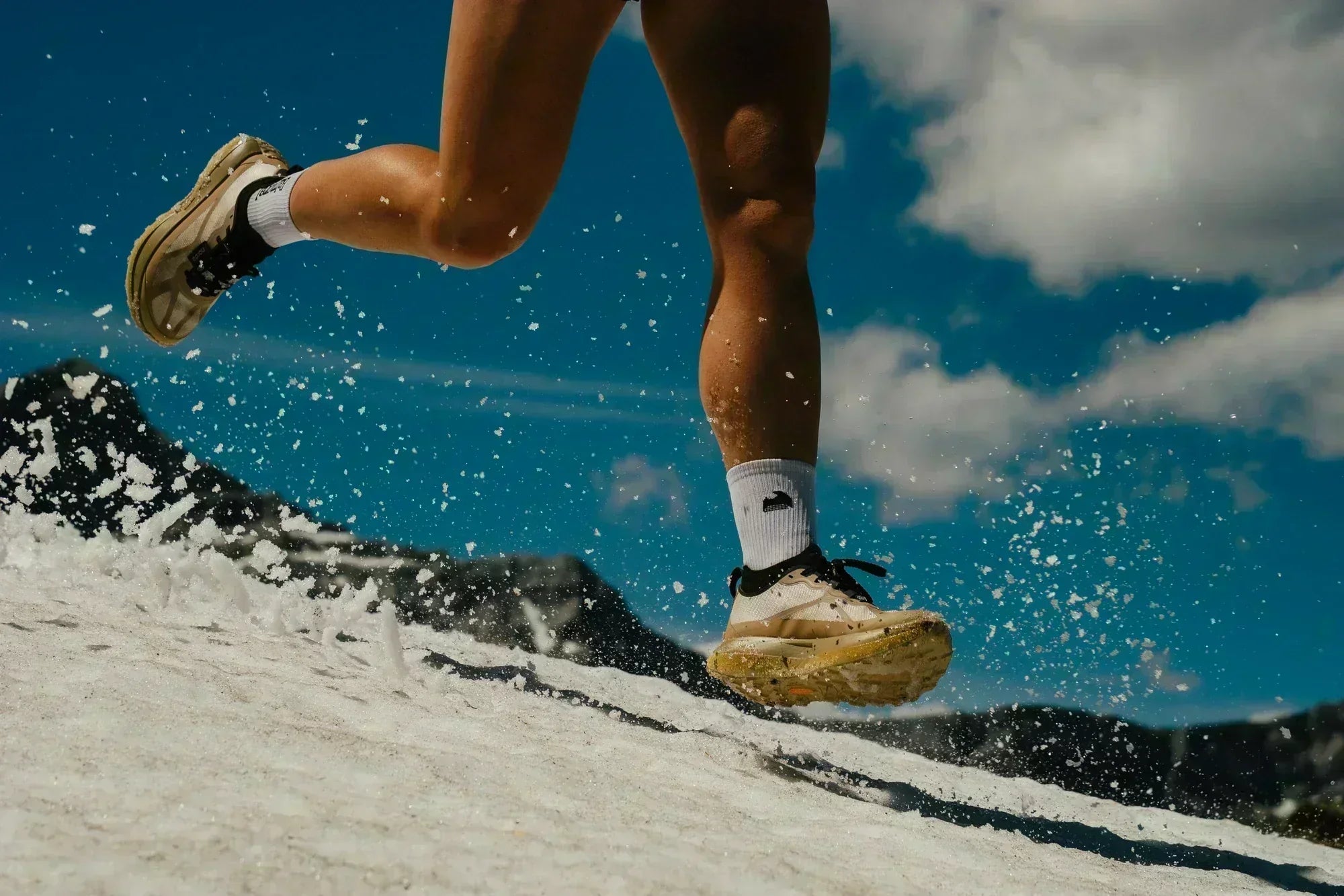 Runner wearing Norda 055 trail running shoes crossing an alpine snowfield with snow spraying beneath each stride.