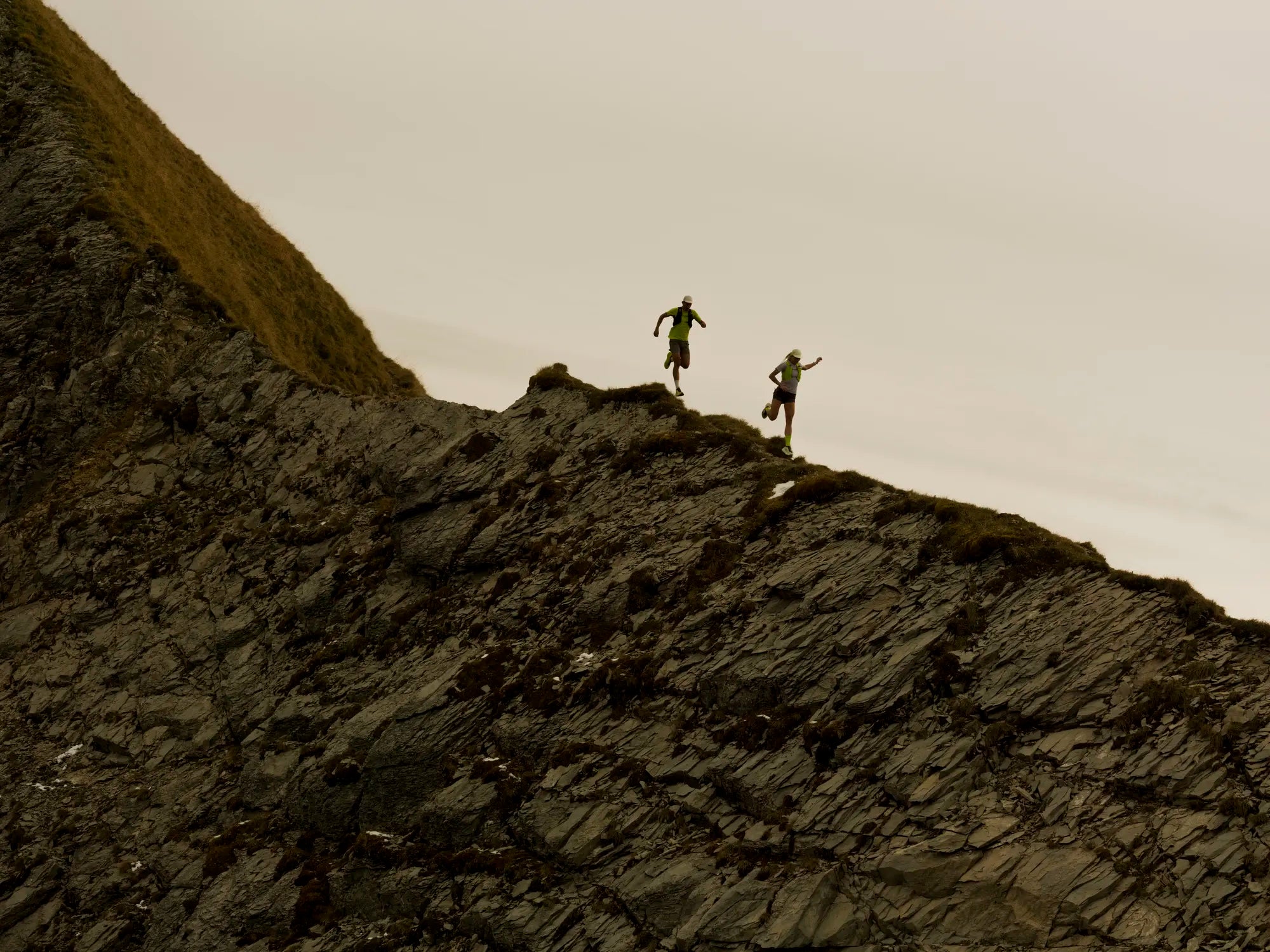 Two people trail running on a rocky ridge with a mountainous landscape.