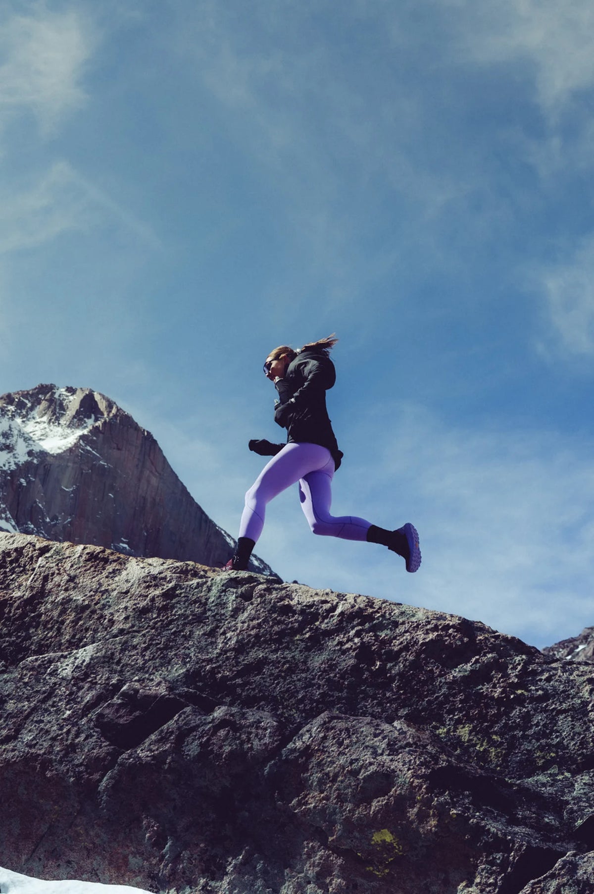A trail runner wearing a black jacket and purple ants running up a rocky ridge.