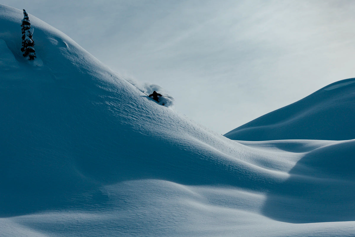 A skier carving through deep powder snow on a dramatic blue-toned mountain slope under a pale sky.