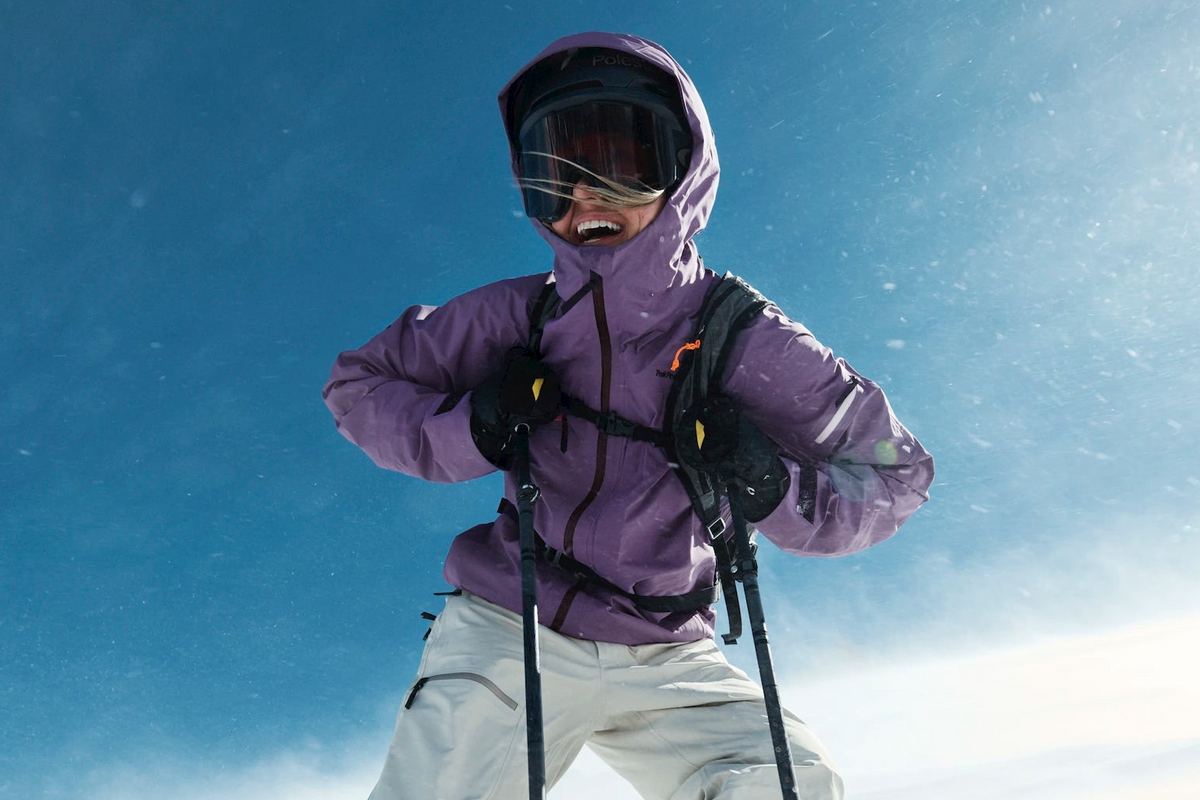 A skier wearing a purple ski jacket, white ski pants, black gloves, and a helmet with goggles, holding ski poles against a bright blue snowy sky.