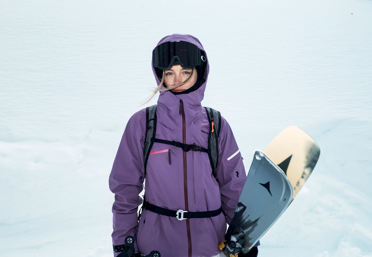 A woman in a purple ski jacket and black goggles holds skis in a snowy outdoor setting.