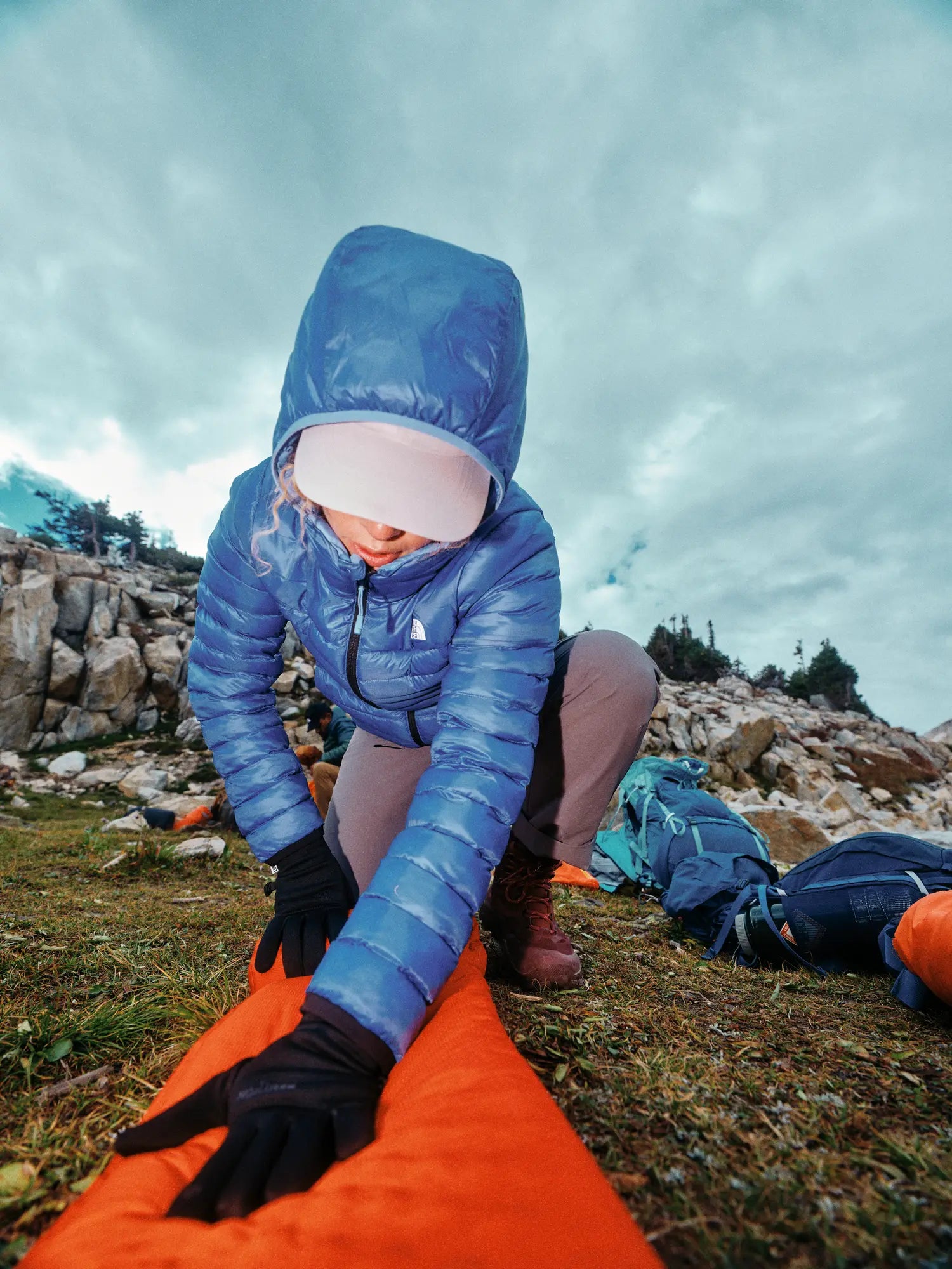 Person in blue jacket setting up an orange sleeping pad on a rocky landscape