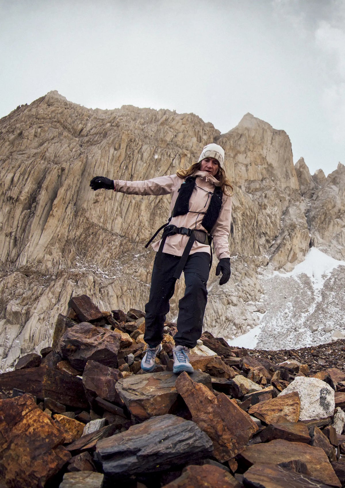 A hiker wearing a pink jacket and black pants walking down loose rocks with mountains in the backdrop.