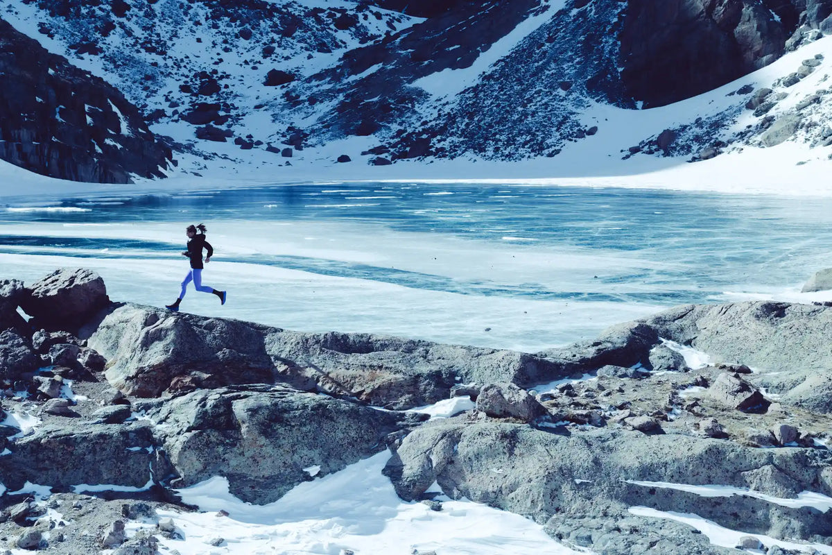 A runner wearing black jacket and purple pants running along a rocky ridge near a frozen over lake with mountains surrounding.