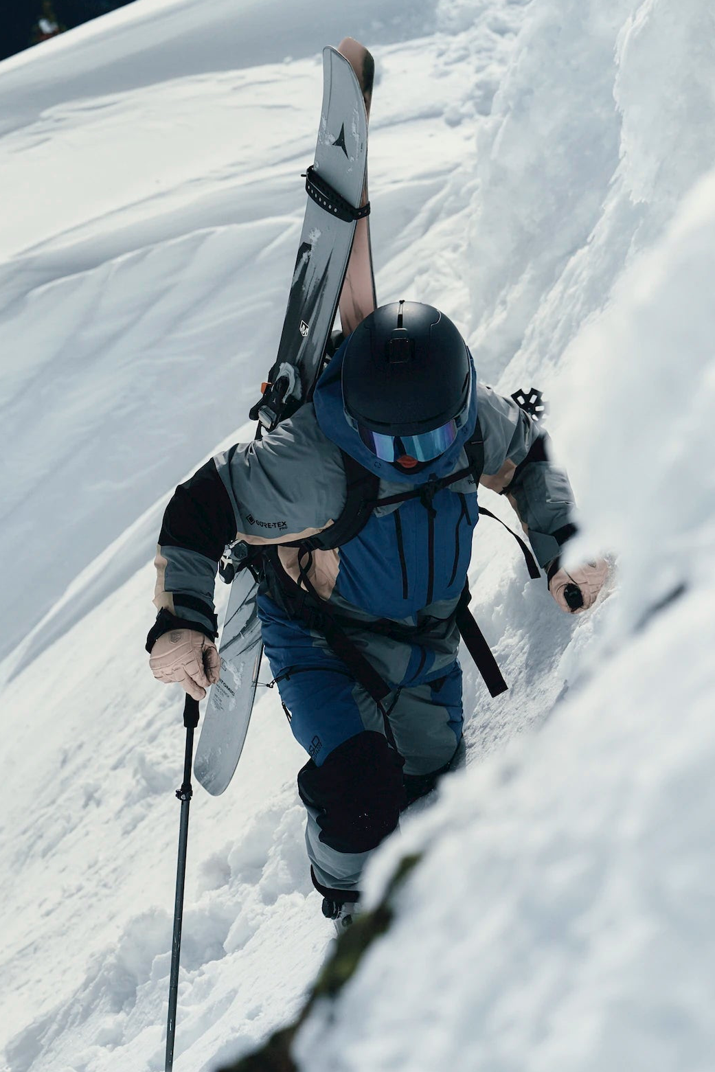 A skier in a gray and blue ski suit with a black helmet and goggles, carrying a white ski up a steep snowy slope.