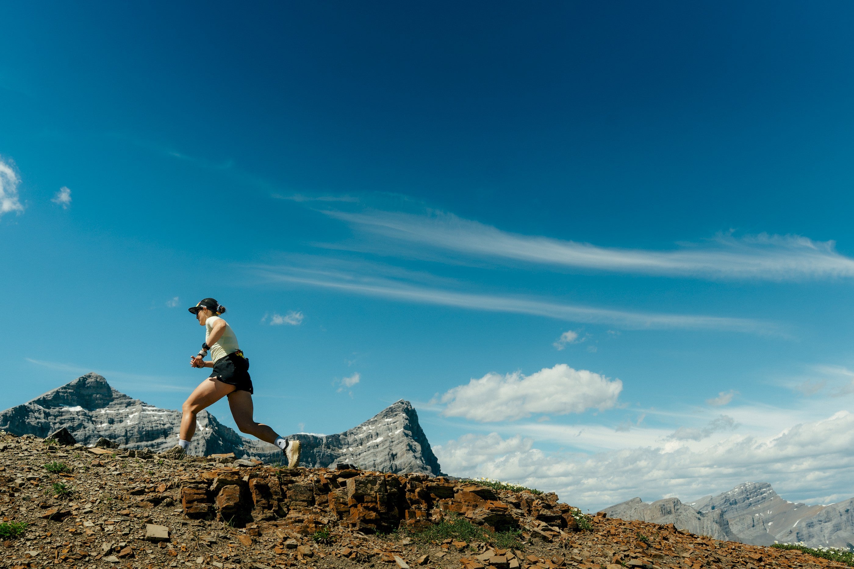 A trail runner in athletic gear sprints up a rocky mountain path with snow-capped peaks and a deep blue sky in the background.