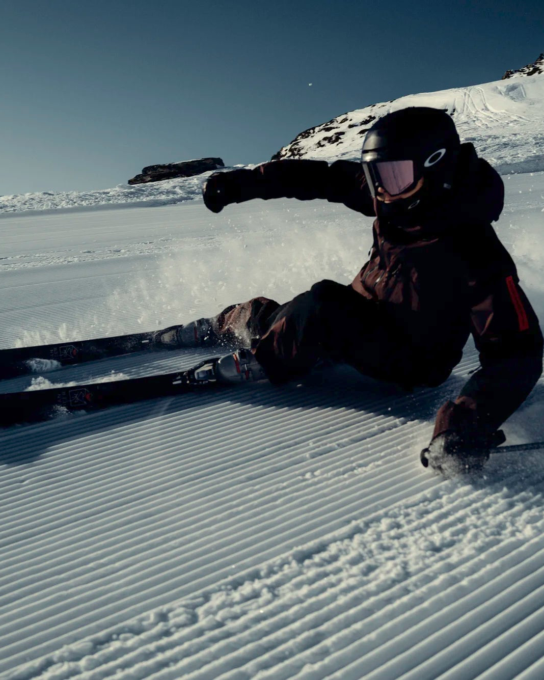 A skier carving down a groomed run with mountains and blue skies in the backdrop.