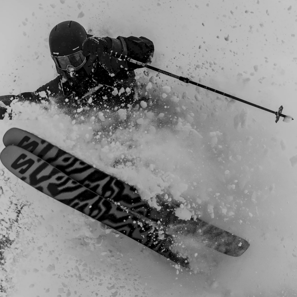 A skier carving through deep powder snow, viewed from above at an angle. The skier wears a dark helmet, goggles, jacket, and gloves, with ski poles and skis visible.
