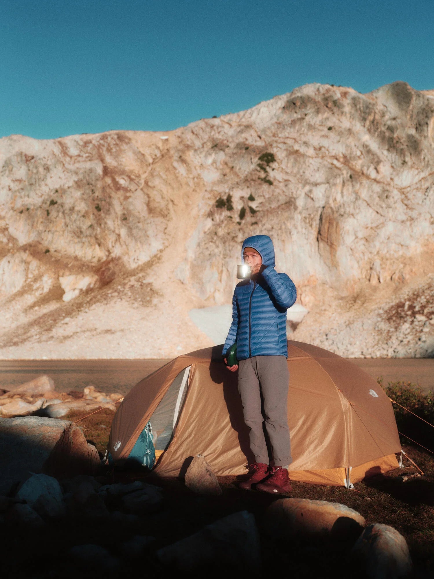 A person drinking from a mug in front of their camp site in a mountain landscape